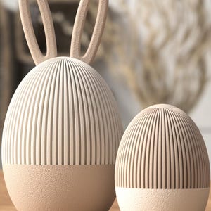 Könnte beinhalten: Zwei beige dekorative, eiförmige Objekte. Eines hat Hasenohren. Beide haben einen strukturierten Sockel und vertikale Linien auf dem oberen Teil. Die Objekte stehen auf einer Holzoberfläche mit verschwommenem Hintergrund.
