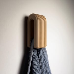 Könnte beinhalten: Ein hellbrauner, U-förmiger Wandhaken mit vertikalen Rillen, der ein blaues Handtuch hält. Der Haken ist an einer hellen Wand befestigt. Das Handtuch hat ein strukturiertes, gemustertes Design.