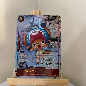 May include: A collectible trading card featuring Tony Tony Chopper from the anime series One Piece. The card has a colorful design with a cartoon illustration of Chopper, text, and a counter. The card is displayed on a small wooden easel.