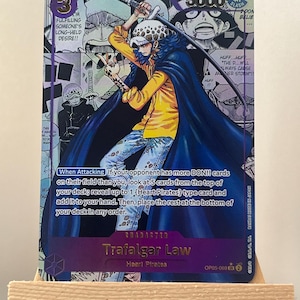 May include: A trading card featuring Trafalgar Law from the Heart Pirates. The card has a purple background with manga-style illustrations and text. Trafalgar Law is depicted in a yellow shirt and blue cape, holding a sword. The card also includes game instructions.