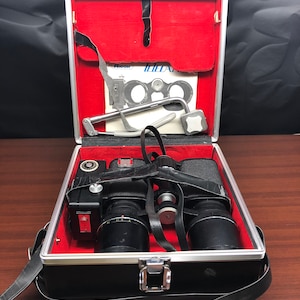 Puede incluir: Cámara negra vintage con dos lentes y una correa, dentro de un estuche plateado y negro con interior rojo. El estuche también contiene un manual y otros accesorios. El estuche está abierto, revelando la cámara y las lentes.