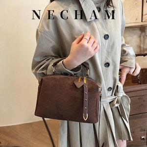 Puede incluir: Un bolso de mano de ante marr&oacute;n con herrajes dorados, incluyendo cremallera y enganches para el asa. El bolso tiene una peque&ntilde;a etiqueta y una correa decorativa. La palabra "NECHAM" est&aacute; impresa en la parte superior.