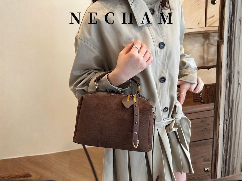 Puede incluir: Un bolso de mano de ante marr&oacute;n con herrajes dorados, incluyendo cremallera y enganches para el asa. El bolso tiene una peque&ntilde;a etiqueta y una correa decorativa. La palabra "NECHAM" est&aacute; impresa en la parte superior.