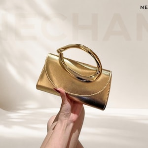 Op de afbeelding: Een metallic gouden handtas met een gladde, gebogen klepsluiting en een opvallend, afgerond, gepolijst goudkleurig metalen handvat. De tas heeft een gestructureerd rechthoekig lichaam met een reflecterende afwerking en de tekst NECHAM erboven.