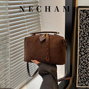 Puede incluir: Un bolso de mano de ante marr&oacute;n con asa superior y bandolera extra&iacute;ble. El bolso presenta una cremallera y herrajes dorados, con la marca "NECHAM" en la parte superior. Una peque&ntilde;a etiqueta de cuero cuelga de la cremallera.