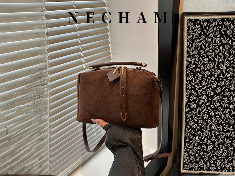 Puede incluir: Un bolso de mano de ante marr&oacute;n con asa superior y bandolera extra&iacute;ble. El bolso presenta una cremallera y herrajes dorados, con la marca "NECHAM" en la parte superior. Una peque&ntilde;a etiqueta de cuero cuelga de la cremallera.