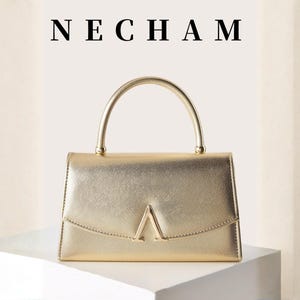 May include: A gold-colored handbag with a curved handle and a geometric design on the front. The word "NECHAM" is displayed above the bag. The bag is sitting on a white surface.