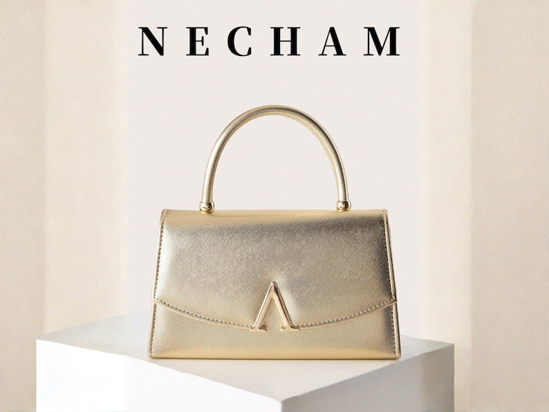 May include: A gold-colored handbag with a curved handle and a geometric design on the front. The word "NECHAM" is displayed above the bag. The bag is sitting on a white surface.