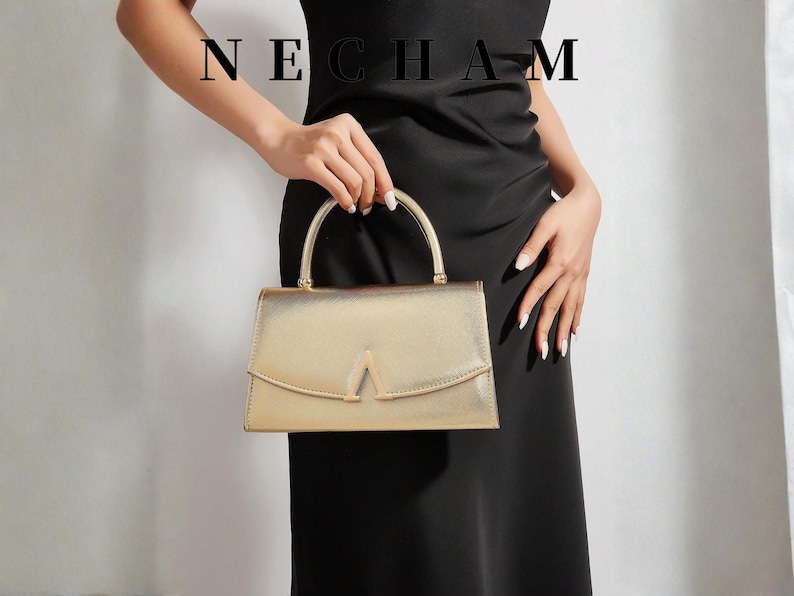 May include: A gold-colored handbag with a structured design and a top handle is held by a person wearing a black dress. The bag features a unique V-shaped detail on the front. The word "NECHAM" is displayed at the top.