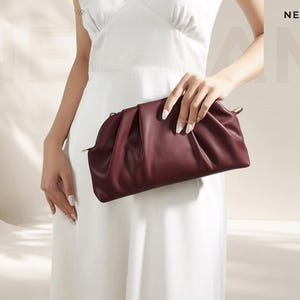 Può includere: Una pochette plissettata color bordeaux intenso con una silhouette morbida e arrotondata e un attacco laterale in metallo dorato. La borsa presenta una finitura liscia e opaca e una struttura rigida, mostrata su sfondo neutro con il testo NECHAM.