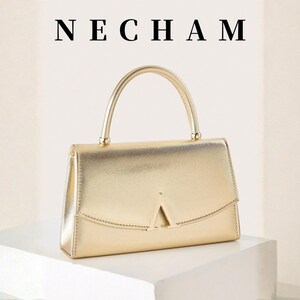 May include: A gold-colored handbag with a top handle and a geometric flap closure. The word "NECHAM" is displayed above the bag. The bag is sitting on a white surface.