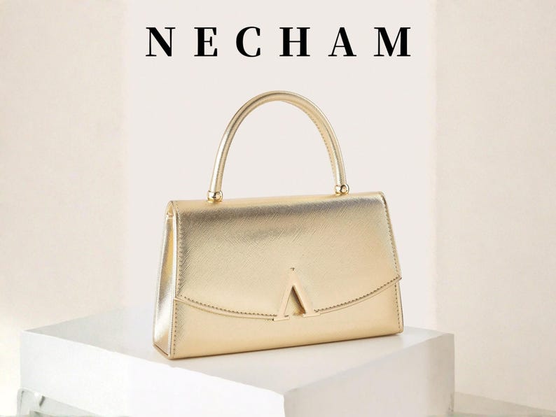 May include: A gold-colored handbag with a top handle and a geometric flap closure. The word "NECHAM" is displayed above the bag. The bag is sitting on a white surface.