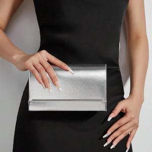 Pode incluir: Clutch prateada com formato retangular e acabamento metálico. A clutch tem um design simples e elegante. É segurada contra um vestido preto.