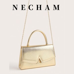 May include: A gold-colored handbag with a top handle and a chain strap. The bag has a rectangular shape with a flap closure and a gold-colored V-shaped detail on the front. The word "NECHAM" is displayed above the bag.