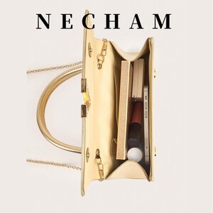 May include: A gold-colored handbag with a top handle and chain strap, open to reveal contents. The bag contains a smartphone, makeup, and other accessories. The word "NECHAM" is printed at the top.