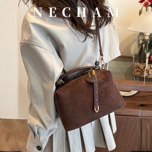 Puede incluir: Bolso de mano de ante marr&oacute;n con asa superior y bandolera extra&iacute;ble. El bolso presenta herrajes dorados y una correa decorativa de cuero con remaches. La palabra "NECHAM" est&aacute; impresa en la parte superior de la imagen.