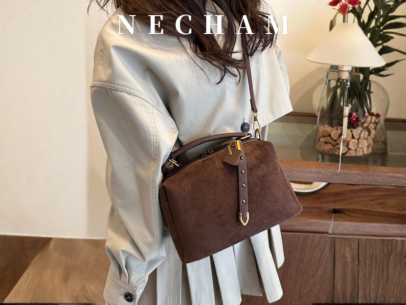 Puede incluir: Bolso de mano de ante marr&oacute;n con asa superior y bandolera extra&iacute;ble. El bolso presenta herrajes dorados y una correa decorativa de cuero con remaches. La palabra "NECHAM" est&aacute; impresa en la parte superior de la imagen.