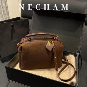 Puede incluir: Un bolso de mano de ante marr&oacute;n con herrajes dorados, incluyendo cremallera y detalles en las asas. El bolso se muestra en una caja negra abierta con una bolsa de la compra a juego en el fondo. La palabra "NECHAM" est&aacute; en la parte superior.