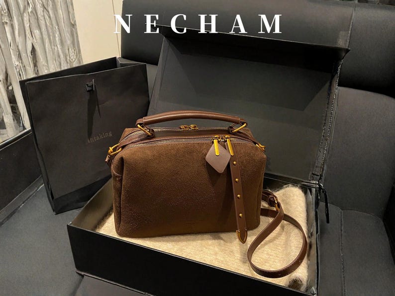 Puede incluir: Un bolso de mano de ante marr&oacute;n con herrajes dorados, incluyendo cremallera y detalles en las asas. El bolso se muestra en una caja negra abierta con una bolsa de la compra a juego en el fondo. La palabra "NECHAM" est&aacute; en la parte superior.