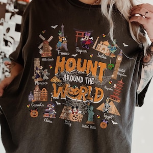 May include: A charcoal gray t-shirt featuring a graphic print with the text "HOUNT AROUND THE WORLD" in orange. The design includes various global landmarks, cartoon characters in costumes, spiderwebs, and pumpkins, representing countries.