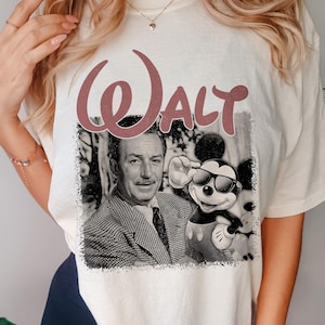 May include: A cream-colored t-shirt featuring a graphic print of Walt Disney and Mickey Mouse wearing sunglasses. The word WALT is printed in large, dusty rose-colored letters above the black and white photographic image of the pair.