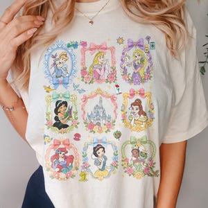 May include: A cream-colored t-shirt featuring a grid of nine ornate, pastel-colored frames. Each frame contains a different illustrated character or a castle, surrounded by floral accents, bows, and small magical icons like a rose and a lamp.