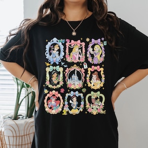 May include: A black t-shirt featuring a graphic print of nine ornate, colorful frames arranged in a grid. Each frame contains a different illustrated character or a castle, decorated with bows, flowers, and small icons like a rose and a lamp.