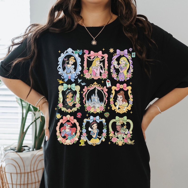 May include: A black t-shirt featuring a graphic print of nine ornate, colorful frames arranged in a grid. Each frame contains a different illustrated character or a castle, decorated with bows, flowers, and small icons like a rose and a lamp.