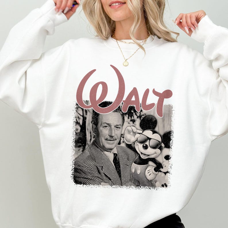 May include: A white long-sleeve sweatshirt featuring a large graphic print on the front. The design includes the word "WALT" in a dusty rose script font above a black and white image of Walt Disney standing next to Mickey Mouse wearing sunglasses.
