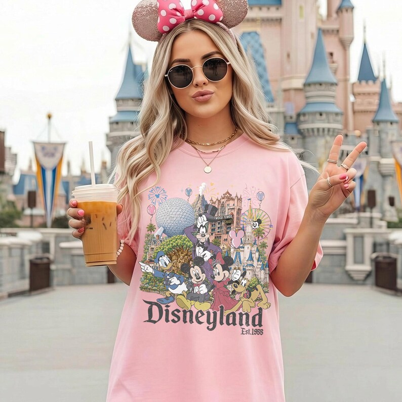 May include: A light pink short-sleeve t-shirt featuring a colorful graphic print of various cartoon characters, a large geodesic sphere, a castle, and the text Disneyland Est. 1955. The shirt has a crew neckline and a relaxed, loose-fitting silhouette.
