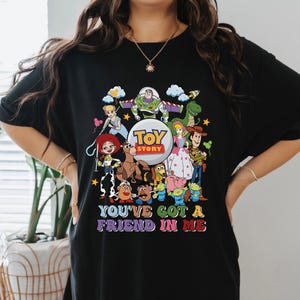 May include: Black t-shirt with a vibrant graphic of characters from the film "Toy Story." The design includes Woody, Buzz Lightyear, Jessie, and others, with the text "You've Got a Friend in Me."