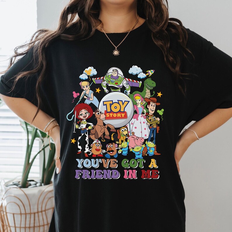 May include: Black t-shirt with a vibrant graphic of characters from the film "Toy Story." The design includes Woody, Buzz Lightyear, Jessie, and others, with the text "You've Got a Friend in Me."