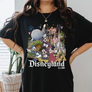 May include: A black t-shirt featuring a colorful graphic print of various cartoon characters including Mickey Mouse, Minnie Mouse, Donald Duck, Goofy, and Pluto in front of a theme park castle, with the text Disneyland Est. 1955 printed below.