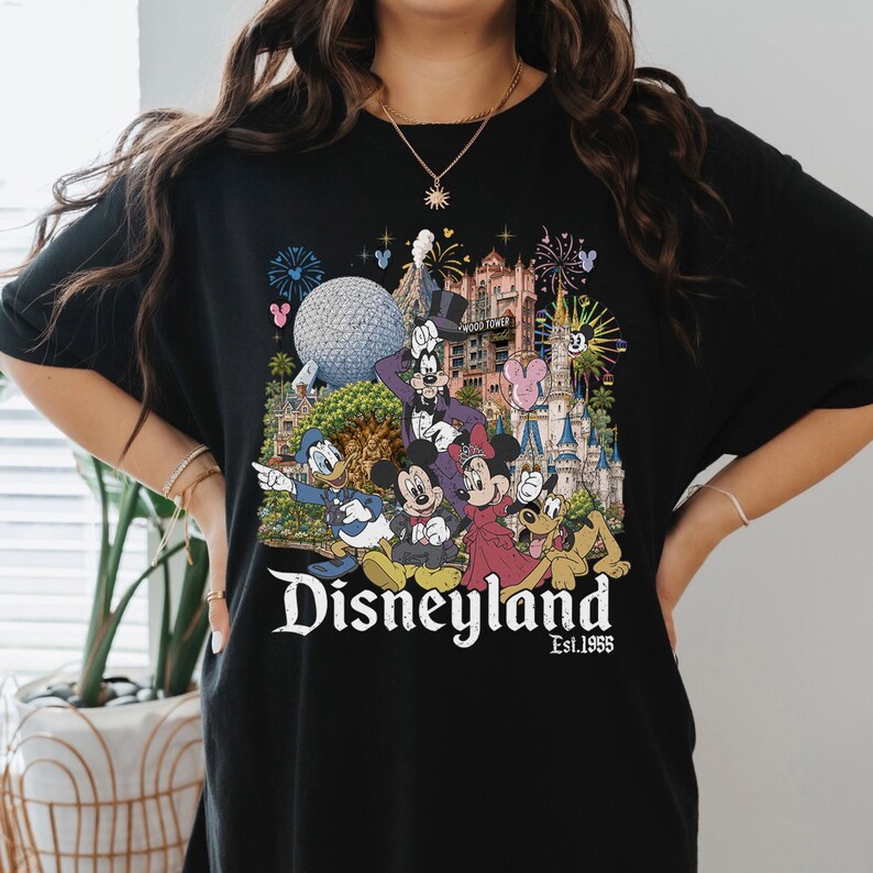 May include: A black t-shirt featuring a colorful graphic print of various cartoon characters including Mickey Mouse, Minnie Mouse, Donald Duck, Goofy, and Pluto in front of a theme park castle, with the text Disneyland Est. 1955 printed below.
