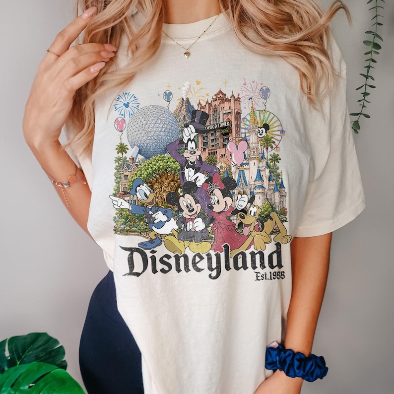May include: A cream-colored t-shirt featuring a colorful graphic of Disney characters including Mickey, Minnie, Donald, Goofy, and Pluto in front of park landmarks. The text reads Disneyland Est. 1955, with fireworks and balloons in the background.