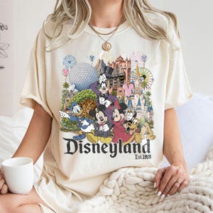 May include: A cream-colored t-shirt featuring a graphic print of various cartoon characters, including Mickey Mouse, Minnie Mouse, Donald Duck, and Goofy, set against a backdrop of theme park landmarks, with the text Disneyland Est. 1955 below.