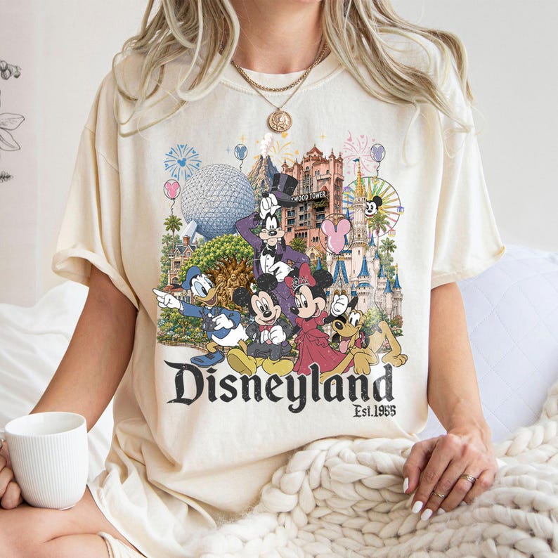 May include: A cream-colored t-shirt featuring a graphic print of various cartoon characters, including Mickey Mouse, Minnie Mouse, Donald Duck, and Goofy, set against a backdrop of theme park landmarks, with the text Disneyland Est. 1955 below.