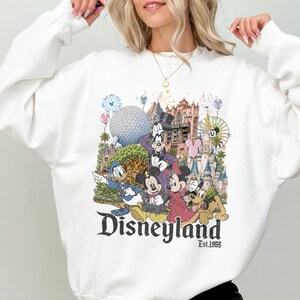 May include: A white long-sleeve sweatshirt featuring a colorful graphic print of Disney characters including Mickey Mouse, Minnie Mouse, Donald Duck, Goofy, and Pluto, with iconic park landmarks and the text Disneyland Est. 1955 printed below.