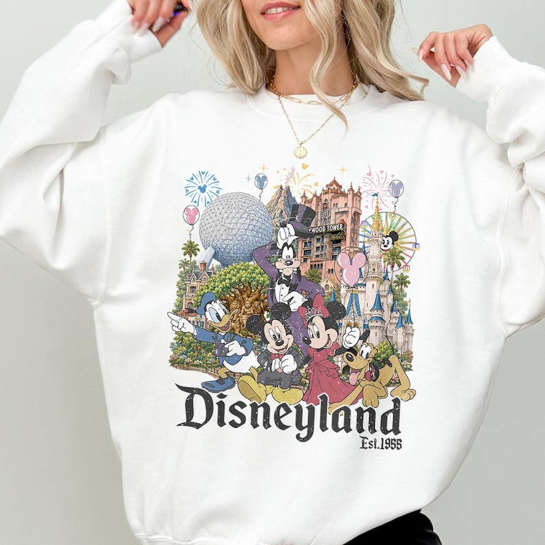 May include: A white long-sleeve sweatshirt featuring a colorful graphic print of Disney characters including Mickey Mouse, Minnie Mouse, Donald Duck, Goofy, and Pluto, with iconic park landmarks and the text Disneyland Est. 1955 printed below.