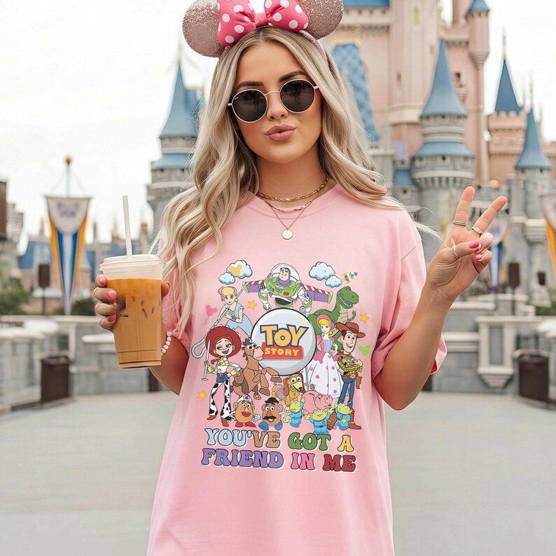May include: A pink t-shirt with a colourful graphic of Toy Story characters, including Buzz Lightyear, Woody, and Jessie. The shirt has the text "Toy Story" and "You've Got a Friend in Me". The person is wearing a pink bow headband and sunglasses.