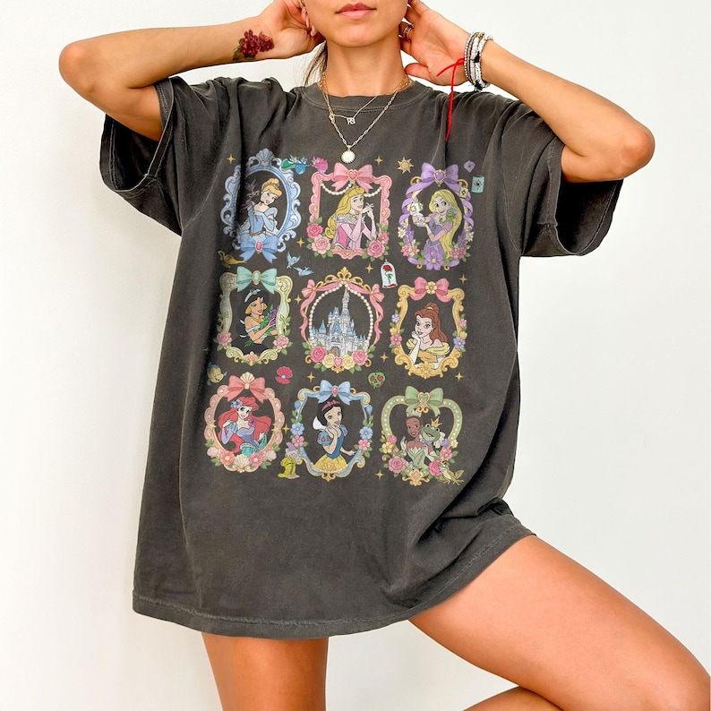 May include: A charcoal gray short-sleeve t-shirt featuring a grid of nine ornate, pastel-colored frames. Each frame contains a different illustrated character or a castle, accented with bows, flowers, and small stars across the front graphic.