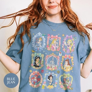 May include: A blue jean colored t-shirt featuring a graphic print of various animated princesses and a castle, each enclosed in ornate, floral-decorated frames. The design includes characters like Cinderella, Aurora, Rapunzel, Jasmine, Belle, Ariel, Snow White, and Tiana.
