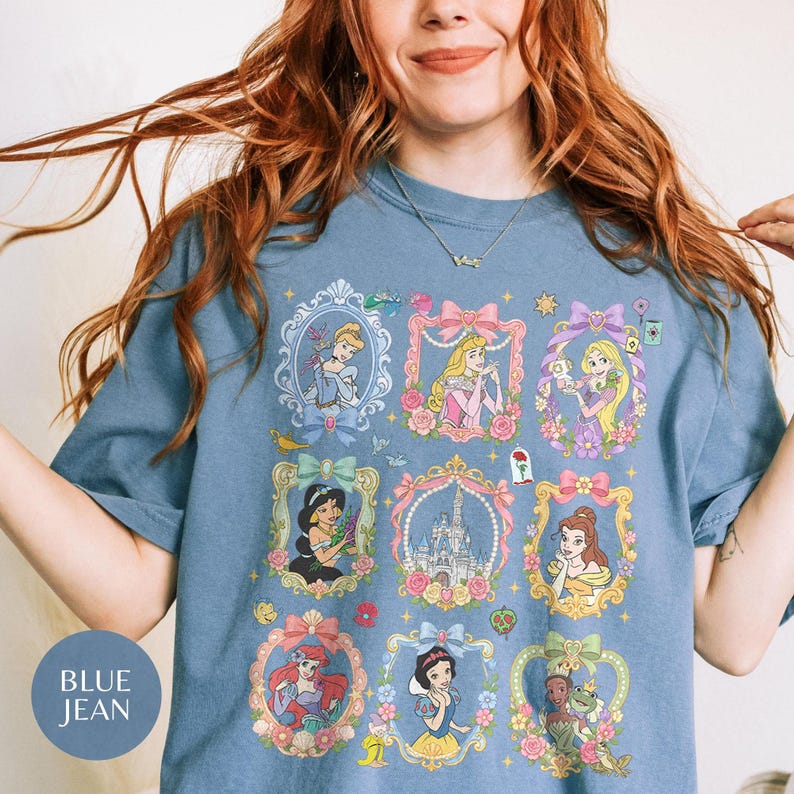 May include: A blue jean colored t-shirt featuring a graphic print of various animated princesses and a castle, each enclosed in ornate, floral-decorated frames. The design includes characters like Cinderella, Aurora, Rapunzel, Jasmine, Belle, Ariel, Snow White, and Tiana.