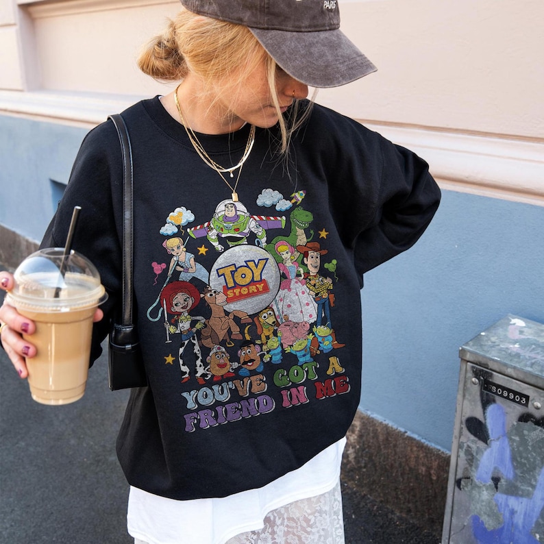 May include: Black sweatshirt with a vibrant Toy Story character graphic. The design showcases Buzz Lightyear, Woody, and other characters from the film, with the text "TOY STORY" and "YOU'VE GOT A FRIEND IN ME".