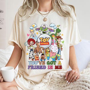 May include: Cream-coloured t-shirt with a vibrant graphic of Toy Story characters, including Buzz Lightyear, Woody, and Jessie. The text "TOY STORY" is in a circle, and the phrase "YOU'VE GOT A FRIEND IN ME" is below the characters.