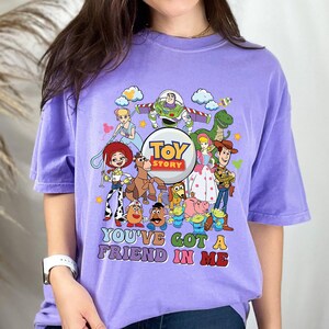 May include: A lilac t-shirt with a vibrant graphic of characters from the animated film "Toy Story." The design includes Buzz Lightyear, Woody, Jessie, and the text "YOU'VE GOT A FRIEND IN ME."