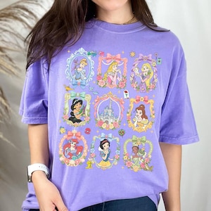 May include: A light purple short-sleeve t-shirt featuring a grid of nine decorative frames. The frames contain illustrations of various animated characters, a castle, and floral accents. The design includes Cinderella, Aurora, Rapunzel, Jasmine, Belle, Ariel, Snow White, and Tiana.
