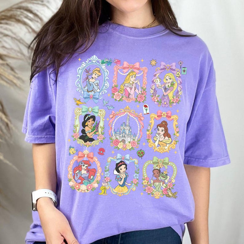 May include: A light purple short-sleeve t-shirt featuring a grid of nine decorative frames. The frames contain illustrations of various animated characters, a castle, and floral accents. The design includes Cinderella, Aurora, Rapunzel, Jasmine, Belle, Ariel, Snow White, and Tiana.