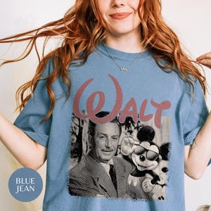 May include: A blue short-sleeve t-shirt featuring a graphic print of Walt Disney and Mickey Mouse wearing sunglasses. The word Walt is printed in a large, stylized pink font above the black and white portrait of the two figures.