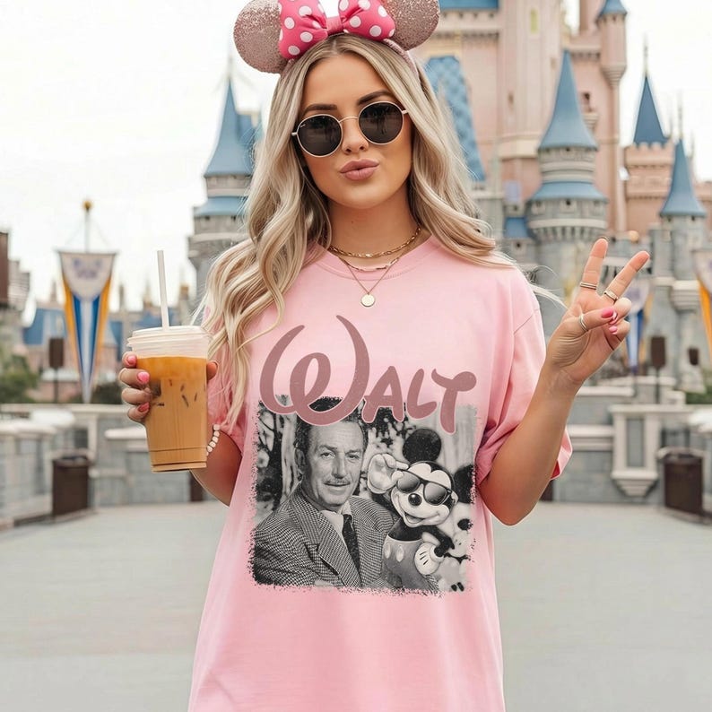 May include: A light pink t-shirt featuring a black and white graphic print of Walt Disney and Mickey Mouse wearing sunglasses. The word "WALT" is printed in a large, stylized pink font above the graphic. The shirt has a crew neckline and short sleeves.